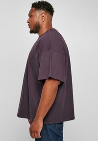 Tricou supradimensionat cu mânecă scurtă, din bumbac purpuriu închis, cu un croi lejer și guler rotund, purtat cu blugi albaștri.