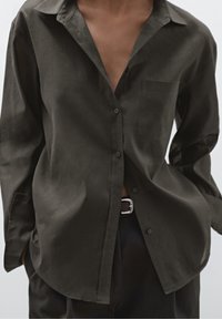 Chemise boutonnée vert foncé avec une texture douce, coupe décontractée, et une poche poitrine. Comprend un col et des manches retroussées.