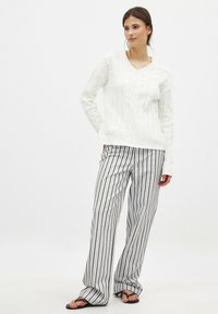 Pull en tricot torsadé blanc avec un col en V, accompagné de pantalons larges à rayures verticales dans des tons de gris et beige. Sandales noires.