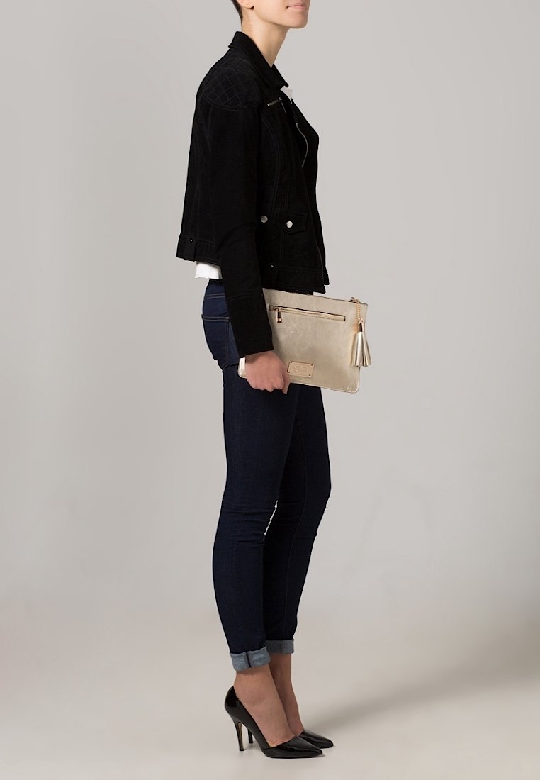 Giacca cropped in suede nera, camicia bianca aderente, jeans scuri e tacchi alti neri. Tiene una clutch beige chiaro con dettaglio a frangia.