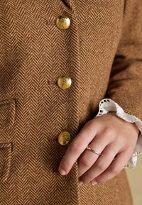 Palton cu model chevron maro și nasturi aurii. Prim-plan cu o mână care ține buzunarul și manseta cu dantelă albă. Material din lână texturată.