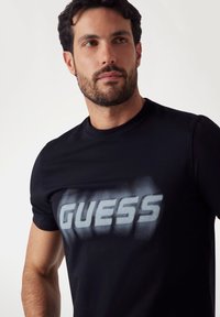 Zwarte katoenen t-shirt met een gedurfde, vervaagde grijze "GUESS" logo op de voorkant. Korte mouwen, ronde halslijn en een gladde textuur.