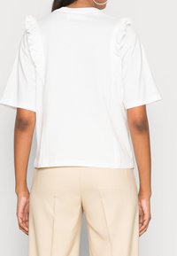 Bílý kratký rukáv top s volánovými akcenty na ramenou, který má volný střih a je vyroben z jemné bavlny. Zobrazeno zezadu.