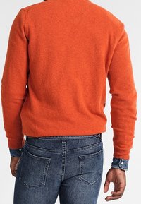 Orange stickad tröja med rund halsringning, bärs över mörk denimjeans. Tröjan har en mjuk textur och ribbat avslut.