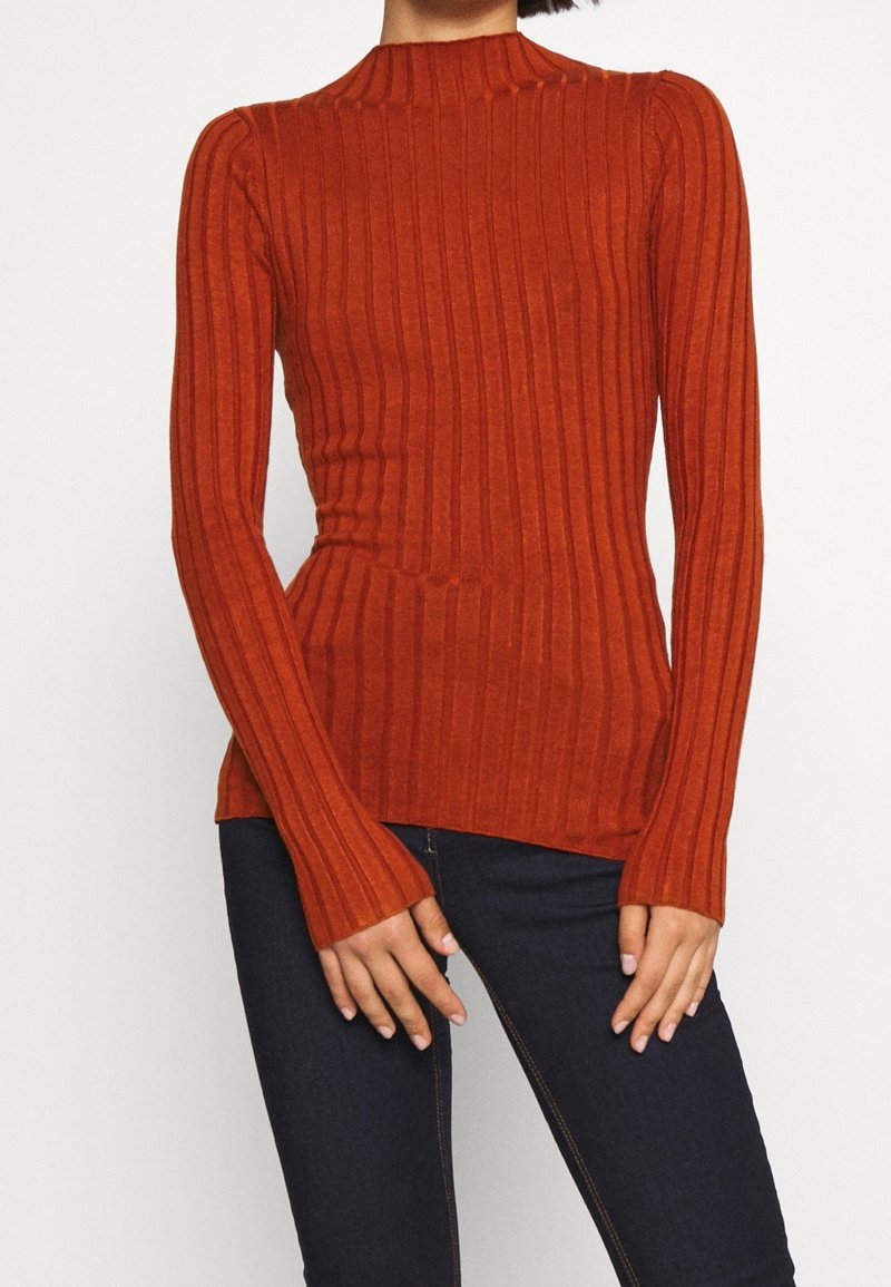 Femme portant un pull ajusté, côtelé, à manches longues de couleur orange rouille et un jean bleu foncé, debout, les mains près des hanches.