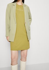 Grüner karierter Blazer über einem senffarbenen Kleid. Der Blazer hat zwei Fronttaschen und einen einzelnen Knopfverschluss. Schlichte kniehohe cremefarbene Stiefel.