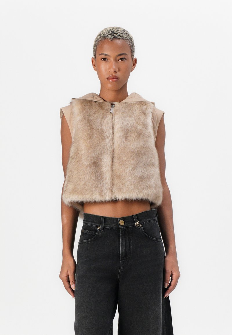Gilet corto in pelliccia sintetica beige con cappuccio, chiusura frontale con zip e senza maniche; indossato sopra jeans scuri a vita alta. Texture liscia e aspetto morbido.