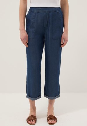 Mujer vistiendo pantalones vaqueros cortos de color azul oscuro con puños enrollados, bolsillos delanteros, camiseta blanca y sandalias marrones tejidas.