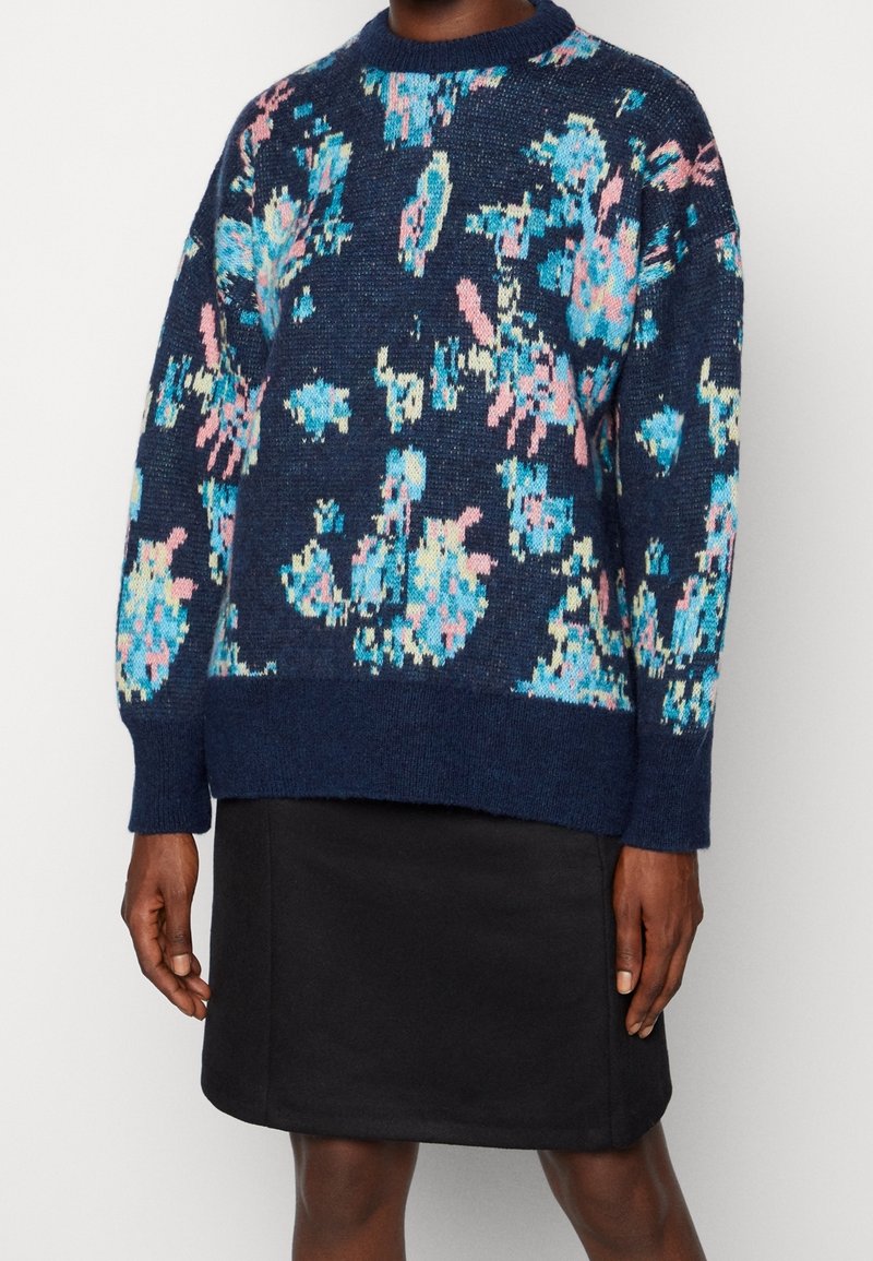 Granatowy sweter dzianinowy z kolorowym abstrakcyjnym wzorem w odcieniach niebieskiego i różowego, okrągły dekolt, luźny krój, zestawiony z czarną spódnicą.