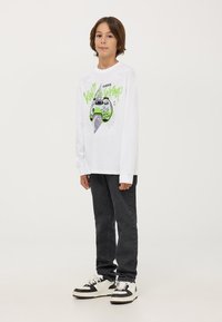 Camicia bianca a maniche lunghe con una grafica di un controller di videogioco verde e testo, abbinata a jeans neri e sneakers in bianco e nero.