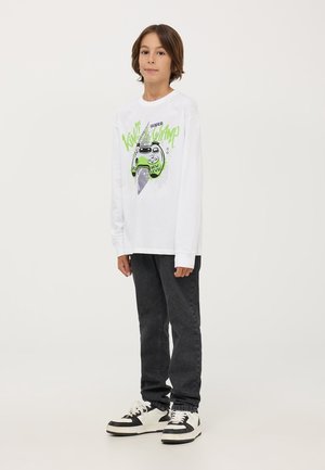 Camicia bianca a maniche lunghe con una grafica di un controller di videogioco verde e testo, abbinata a jeans neri e sneakers in bianco e nero.