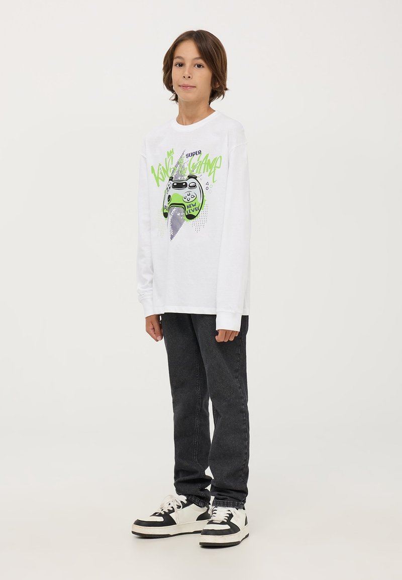 Camicia bianca a maniche lunghe con una grafica di un controller di videogioco verde e testo, abbinata a jeans neri e sneakers in bianco e nero.