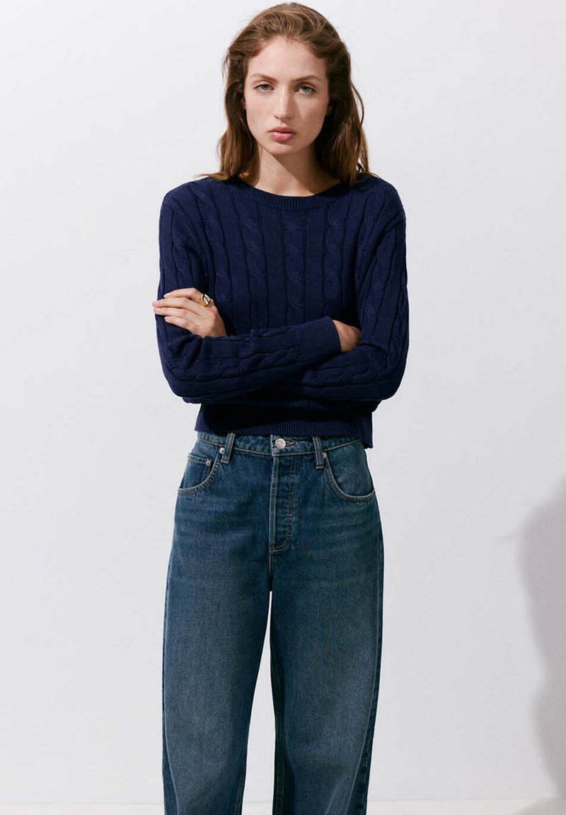 Femme aux cheveux châtain clair, debout les bras croisés, portant un pull torsadé bleu marine et un jean bleu taille haute sur fond blanc.