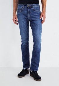 Vaqueros de mezclilla en un tono azul medio, corte recto, con desgastado ligero, cinco bolsillos y cierre de botón. Combinados con zapatillas negras.