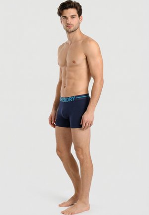 Jeune homme debout pieds nus en boxer bleu marine avec ceinture « Superdry » sur fond gris clair.