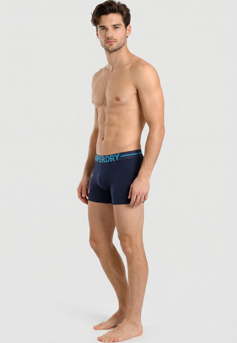 Jeune homme debout pieds nus en boxer bleu marine avec ceinture « Superdry » sur fond gris clair.