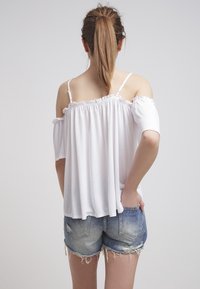 Mladá žena s hnědým culíkem, oblečená v bílé off-shoulder blůze a roztrhaných denimových šortkách, stojící s jednou rukou v kapse, zády k divákovi.