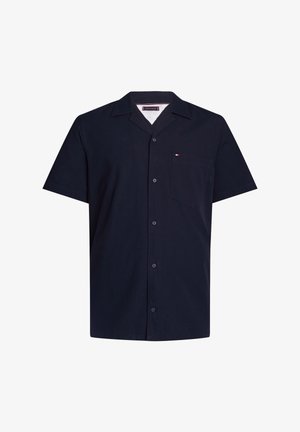 Camisa de manga corta azul marino con botones, cuello puntiagudo y bolsillo en el pecho con un pequeño logo en rojo, blanco y azul.