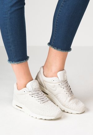Osoba w niebieskich dżinsach z postrzępionymi nogawkami i białych butach Nike Air Max z fakturowanym wzorem, stoi na jasnej podłodze.