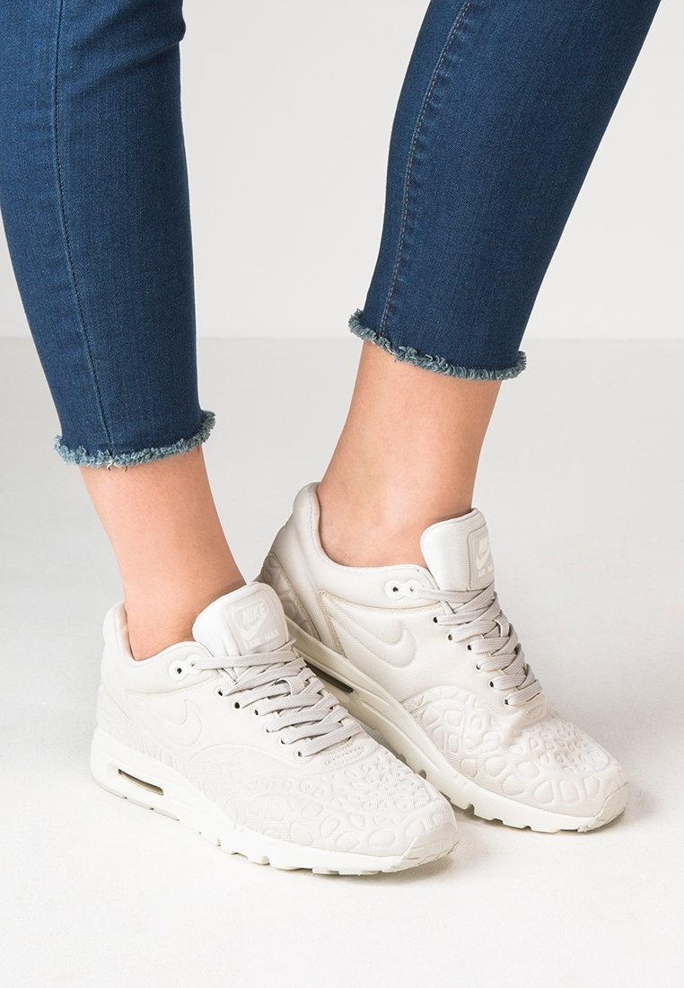 Personne portant un jean bleu à ourlet effiloché et des baskets Nike Air Max blanches à motif texturé, debout sur un sol clair.