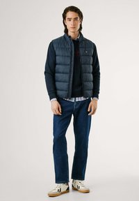 Marineblå puffer vest over en mørk genser, kombinert med blå jeans og kremfargede sneakers. Vestet har høy krage og glidelås foran.