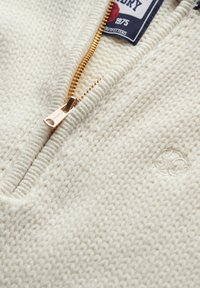 Pull en maille texturée couleur crème avec fermeture éclair. Il présente une fermeture éclair dorée et un logo brodé près de l'ourlet.