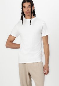 T-shirt branco de algodão com um clássico decote redondo, mangas curtas e uma silhueta ajustada, combinado com calças bege claras.