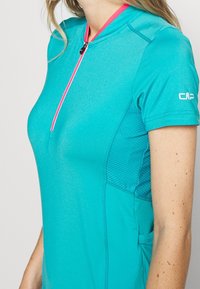Camiseta atlética de manga corta en color teal con una cremallera rosa, paneles laterales de malla y un logotipo en la manga. Textura suave y diseño ajustado.