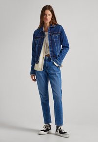 Mörkblå denimjacka med knäppning fram, två bröstfickor och lätt slitna kanter, ihop med ljusblå jeans och svarta sneakers.