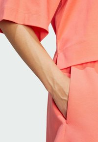 Koraliowy top z bawełny z krótkim rękawem, charakteryzujący się luźnym krojem i boczną kieszenią, podkreślający gładką fakturę i prosty design.