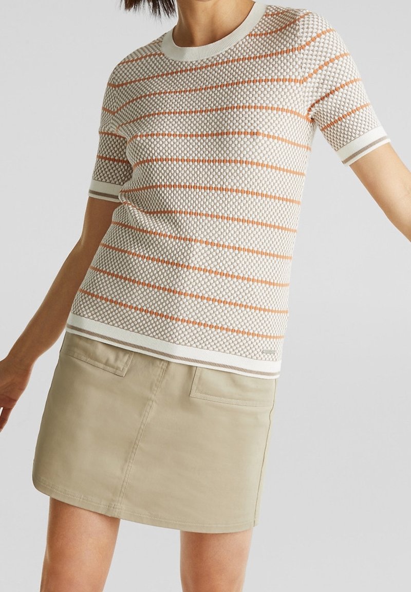 Femme portant un haut en maille texturée à manches courtes avec des rayures orange et une mini-jupe beige avec des poches à l'avant.