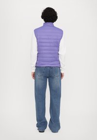 Colete roxo puff com gola alta, usado sobre uma camisa branca de mangas compridas. Combinado com jeans azuis largos e sapatos pretos.