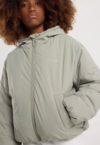 Giacca imbottita verde chiaro con cappuccio, dotata di chiusura frontale con zip, tasche laterali e un logo discreto sul petto. Realizzata in tessuto morbido e liscio.