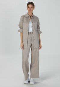Hellbeige Leinenhemd mit Knopfleiste, langen hochgekrempelten Ärmeln, kombiniert mit passender weit geschnittener Hose. Weiße Sneakers runden den Look ab.