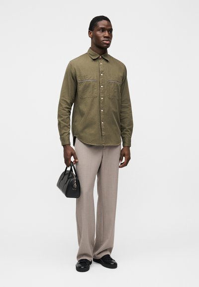 Πουκάμισο χακί με κουμπιά και δύο τσέπες στο στήθος με γκρι λεπτομέρειες, συνδυασμένο με ανοιχτό μπεζ παντελόνι και μικρή μαύρη τσάντα.