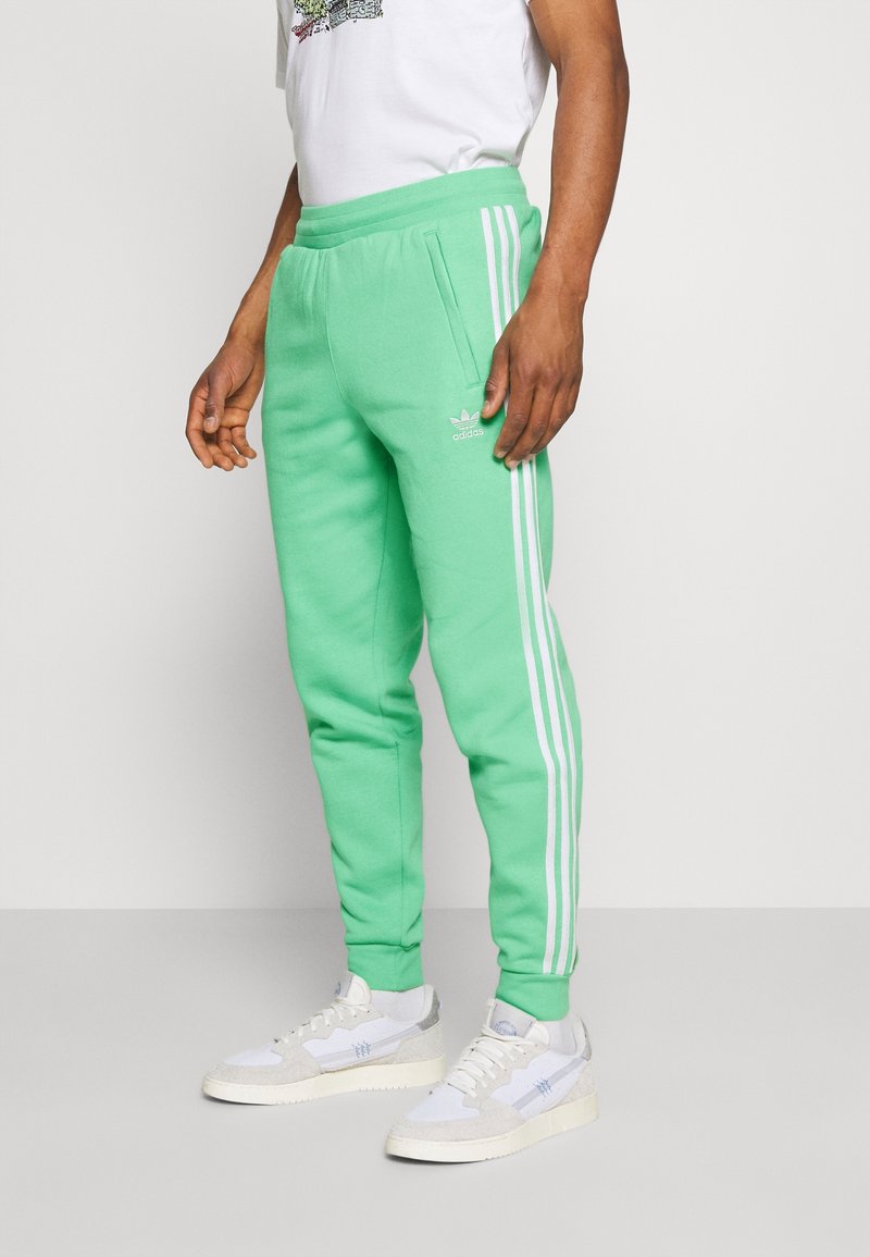 Muž oblečený v jasně zelených teplákových kalhotách Adidas s bílými pruhy, bílých teniskách a bílém tričku s grafikou, stojící na jednolitém pozadí.
