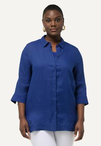 Camisa de lino azul de botón con cuello, mangas tres cuartos y un corte relajado. Combinada con una camiseta azul marino y pantalones blancos.