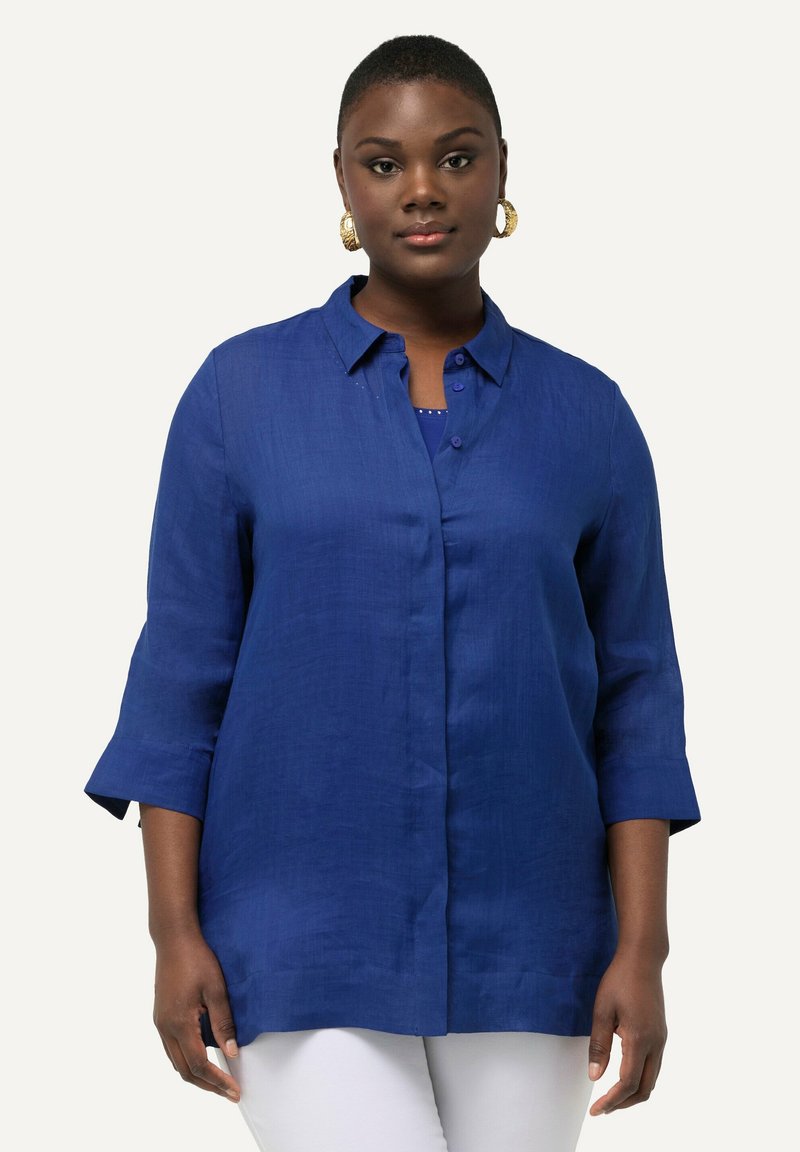 Camisa de lino azul de botón con cuello, mangas tres cuartos y un corte relajado. Combinada con una camiseta azul marino y pantalones blancos.