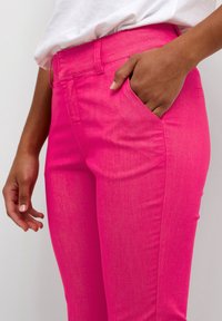 Pantaloni rosa brillante in denim con una texture liscia. Presentano una tasca laterale e passanti per la cintura, vestibilità sagomata e dettagli di cucitura minimi.