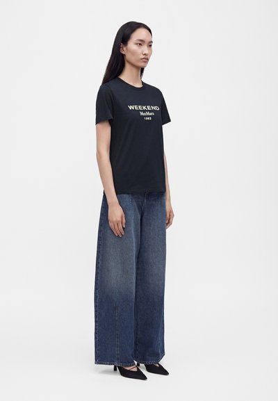 Femme portant un t-shirt noir "WEEKEND Max Mara 1983", un jean bleu délavé à jambes larges et des talons noirs pointus, debout sur un fond blanc.