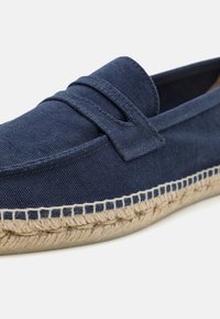 Zapato slip-on de lona azul marino con suela de yute cosida, que presenta una correa decorativa y tela texturizada. Forro suave y construcción flexible.