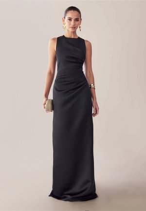 Vrouw in een zwarte avondjurk zonder mouwen, met een beige clutch in haar hand, gouden opvallende oorbellen en armband dragend, tegen een beige achtergrond.