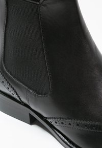 Čern�á kožená kotníková bota s texturovanou elastickou boční částí a dekorativním perforovaným detaily podél podešve. Hladký povrch, robustní podpatek.