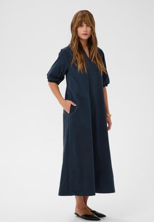 Žena s dlhými hnedými vlasmi, oblečená v námornícky modrých midi šatách s rukávmi do lakťov, s vreckami, čiernymi špicatými balerínkami a zlatým náramkom.