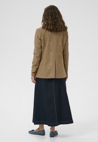 Bruine textuurblazer met geborduurde accenten, gecombineerd met een lange donkerdenimrok en blauwe flats. Het ontwerp heeft een rugventilatie.