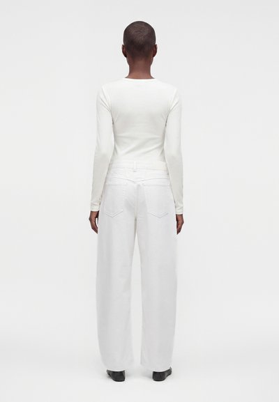 Personne aux cheveux courts portant une chemise blanche à manches longues et un pantalon blanc large, debout de dos sur un fond uni.