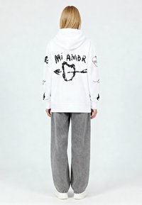 Felpa con cappuccio bianca con grafiche nere, con scritto "Mi Amor" e vari simboli sul retro, abbinata a pantaloni grigi a gamba larga e sneakers bianche.