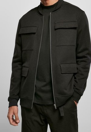 Homme portant une veste noire zippée avec quatre poches à rabat sur le devant et une chemise noire, vu des épaules au milieu des cuisses, mains détendues.