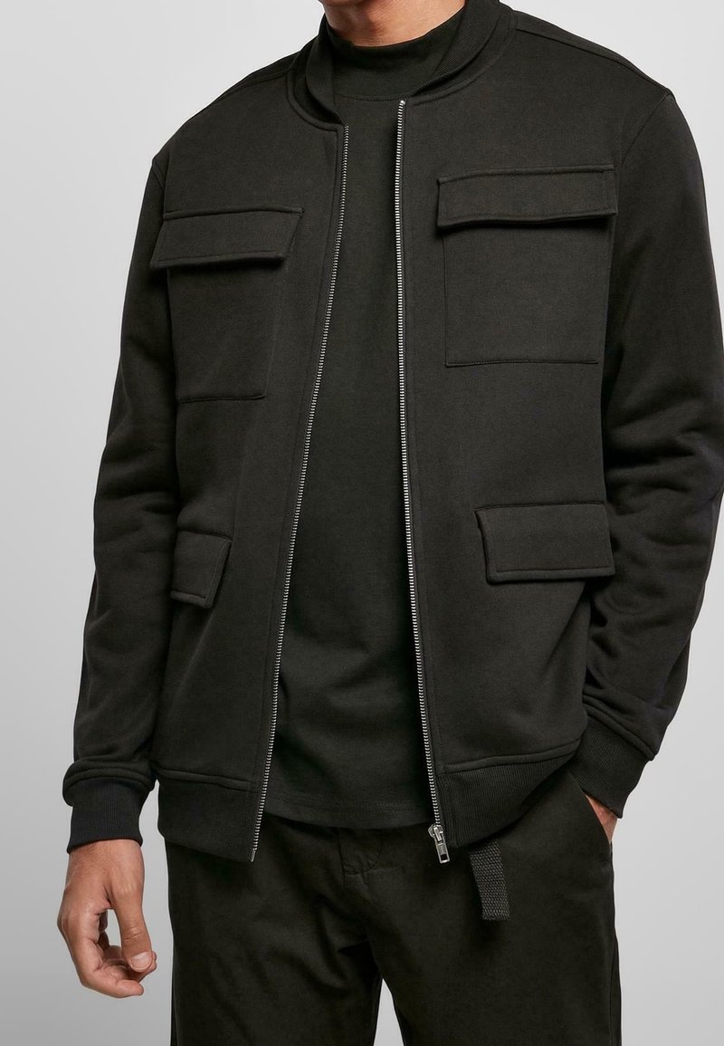 Homme portant une veste noire zippée avec quatre poches à rabat sur le devant et une chemise noire, vu des épaules au milieu des cuisses, mains détendues.