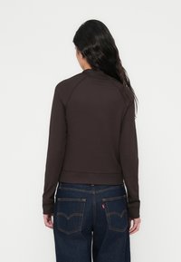 Καφέ, στενή cropped ζακέτα με ριμ π ύφανση και μακριά μανίκια, με ψηλό γιακά και εφαρμοστή μέση, συνδυασμένη με σκούρα μπλε τζιν παντελόνια με πίσω τσέπες.